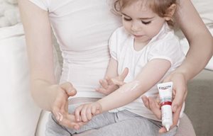 Une petite fille, assise sur les genoux de sa mère, se met de la crème calmante intensive Eucerin AtopiControl dans le pli du coude.